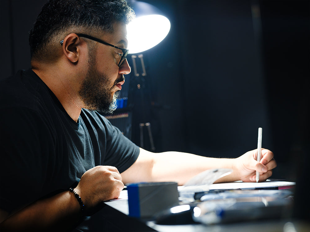 A man with glasses and a beard sits at a desk under a bright lamp, focused on writing or drawing on a tablet with a stylus in a dimly lit room.