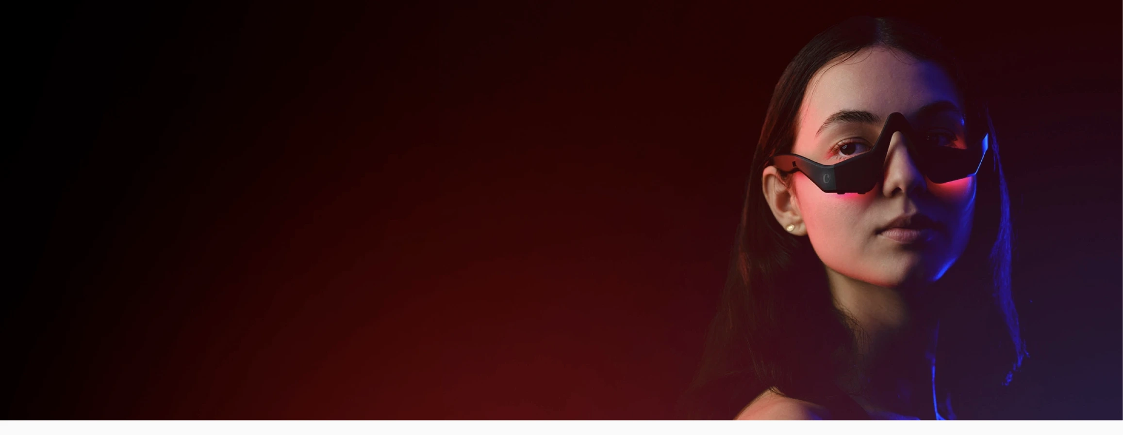 A woman with long dark hair wears futuristic, black and red goggles, gazing confidently forward. She is illuminated by red and blue light, with a dark background.