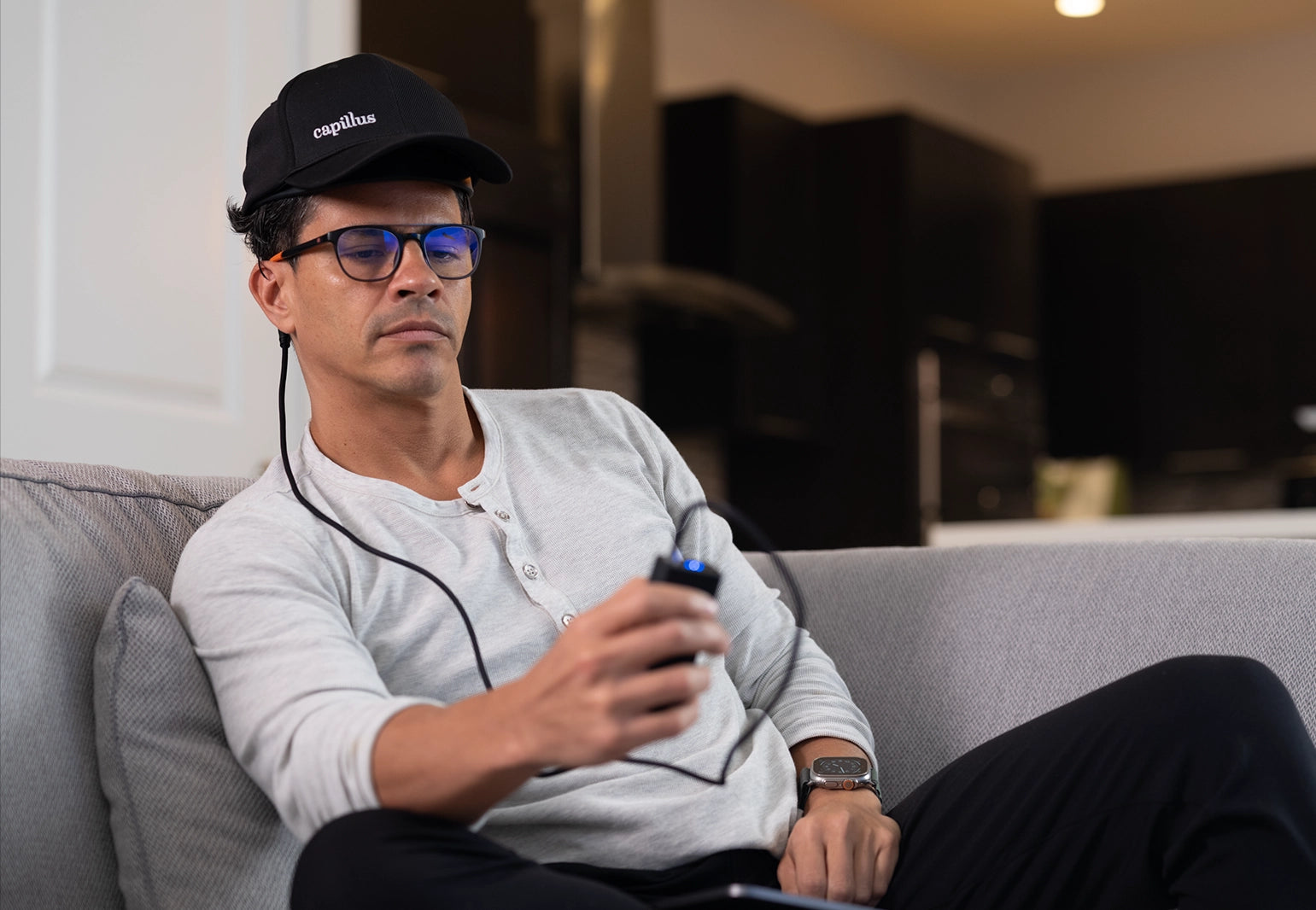 A man wearing glasses and a black cap sits on a couch, holding a small electronic device connected by wires to his cap. He appears focused on the device, and the background shows a modern, dimly lit room.