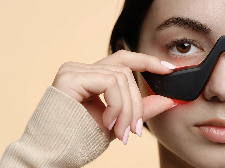 A person with light skin and neatly groomed eyebrows holds futuristic black glasses with red accents close to their eye. They wear a beige sweater, and their nails are manicured in a soft pink color.