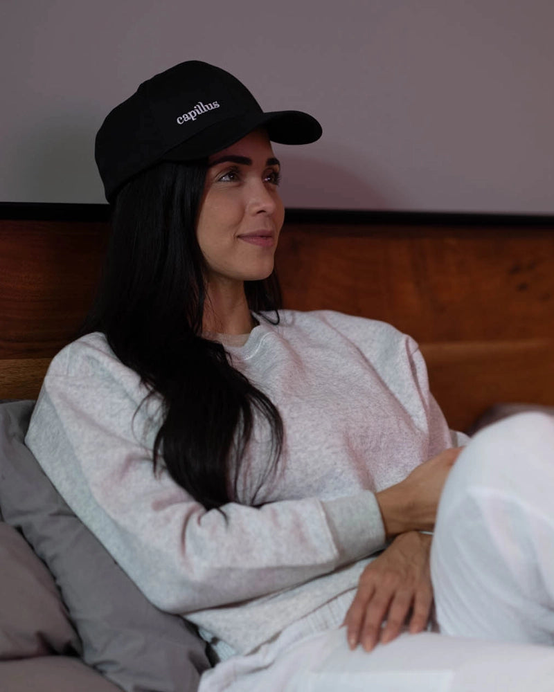 A woman with long dark hair, wearing a black cap and a light gray sweatshirt, sits on a gray couch against a wooden wall, looking to the side and smiling slightly.