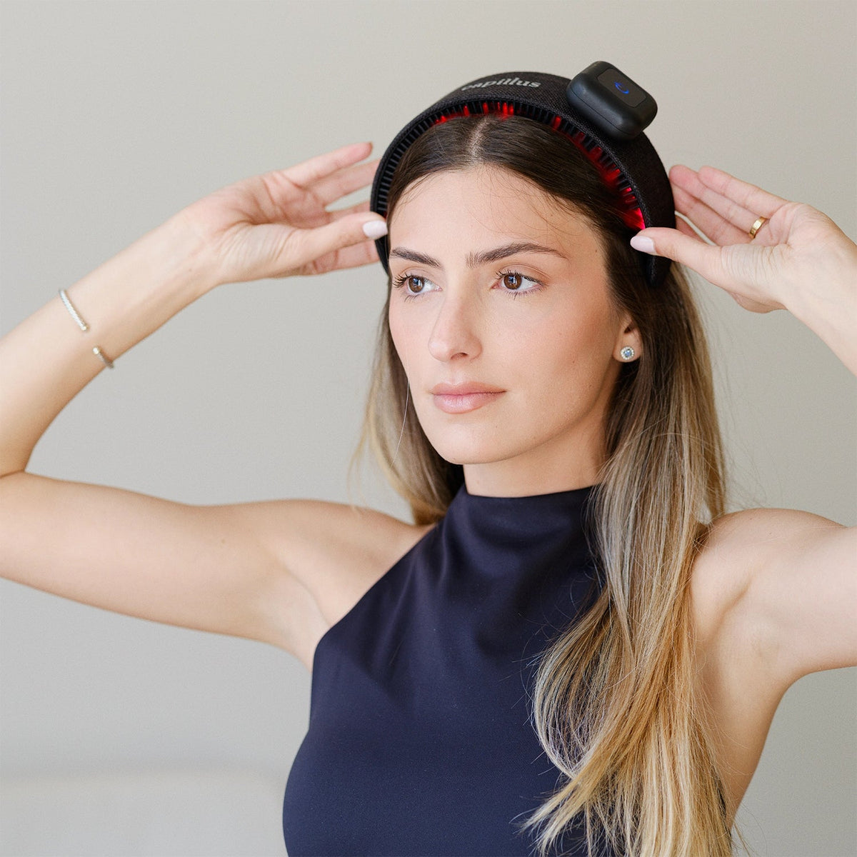 A woman with long hair wears a black sleeveless top and adjusts the Capillus HairBand by Capillus—a state-of-the-art hair wellness wearable equipped with red medical-grade laser technology—on her head. She appears focused, set against a neutral background.