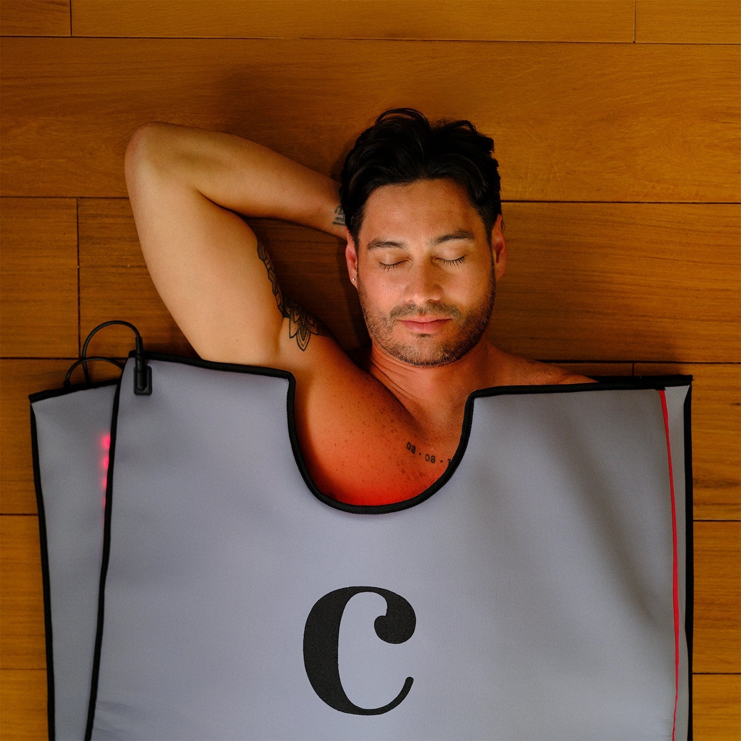 A man with dark hair relaxes with his eyes closed inside the gray Capillus Healing Pod sauna blanket by Capillus, featuring a large black C on the front—ideal for muscle recovery—set against a wooden wall background.