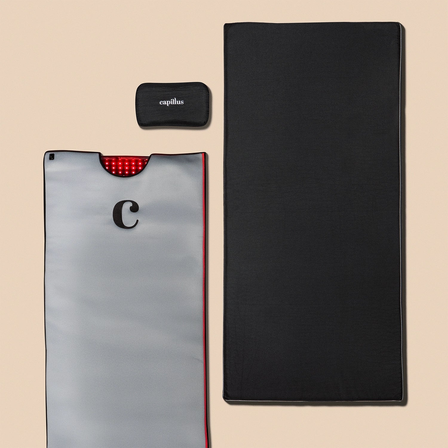 The Capillus Healing Pod by Capillus, a gray rectangular mat featuring a prominent "C" and black mask, specially designed for red light therapy, is displayed next to a plain black rectangular mat on a beige background. This setup is intended to support joint pain relief and muscle recovery.