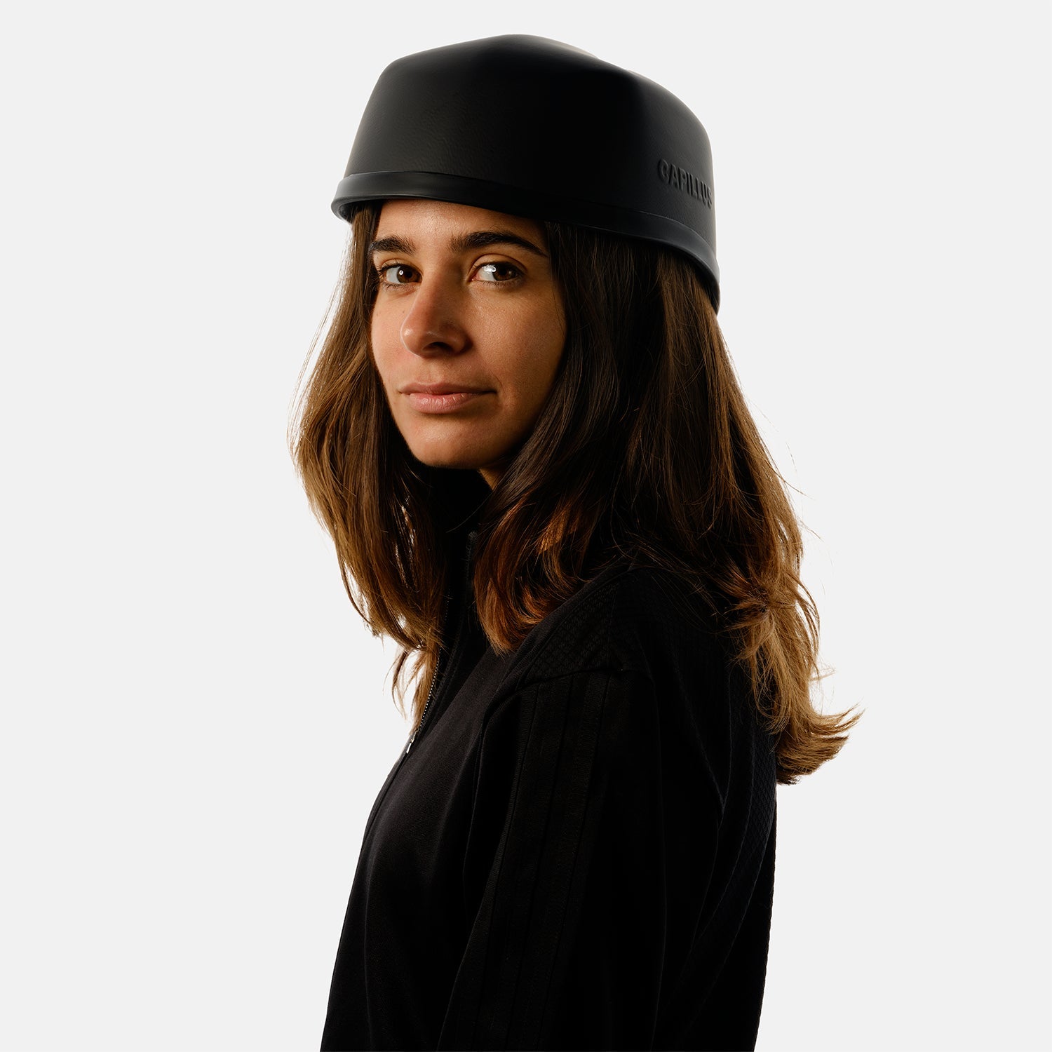 Une femme aux longs cheveux bruns porte un Capillus Neuro de Capillus — ce casque discret, de couleur noir mat, favorise les performances cognitives grâce à un système de photobiomodulation —, assorti d'une veste noire ; elle se tient debout devant un fond blanc et regarde l'appareil photo d'un air neutre.