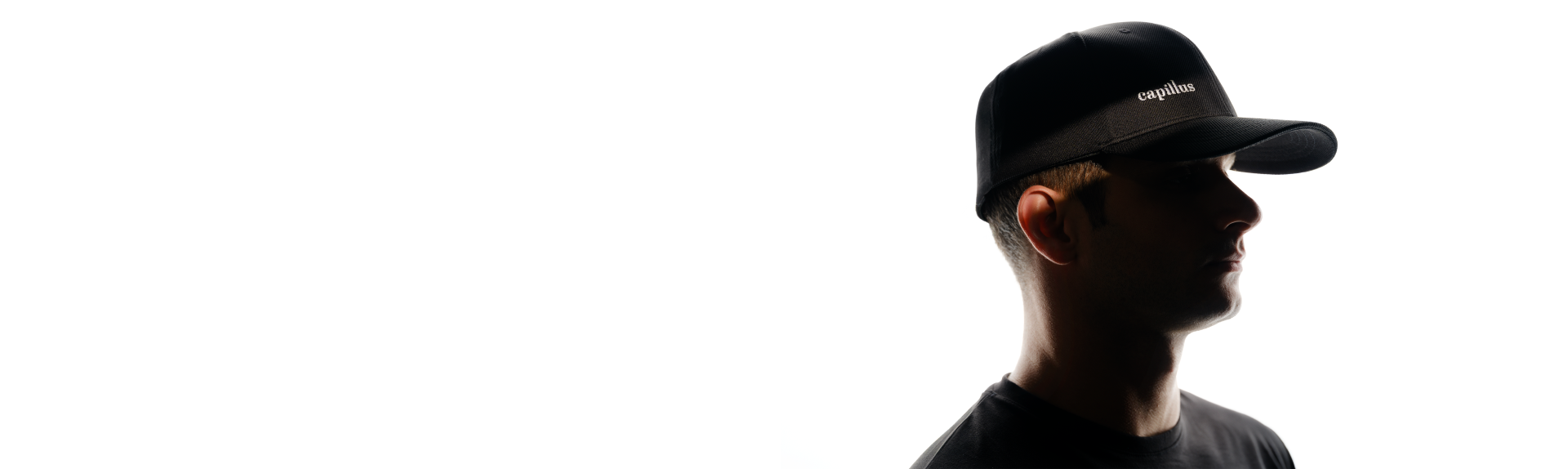 A man in a black shirt and black cap is shown in profile against a white background, with his face mostly in shadow. The cap has text on the front.