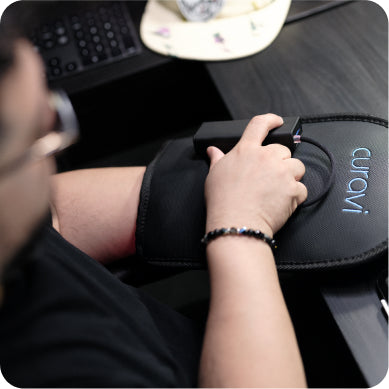 A person using a mouse on a black Auravi wrist rest pad at a desk, with part of a keyboard and a light-colored cap visible in the background.