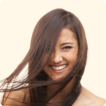 Une femme aux longs cheveux bruns et raides sourit de tout son cœur, quelques mèches lui tombant sur le visage. L'arrière-plan est uni et de couleur claire.