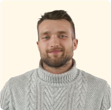 Un homme aux cheveux bruns courts et à la barbe, vêtu d'un pull à col roulé gris clair en maille torsadée, sourit doucement devant un fond clair et uni.