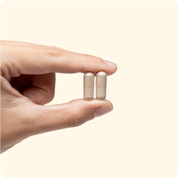 Une main tenant deux gélules beiges entre le pouce et l'index, sur un fond uni et clair.