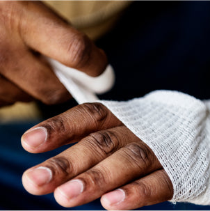 A close-up of a person’s hand being wrapped with a white gauze bandage, suggesting care for an injury. The background is dark, making the hand and bandage the focus of the image.