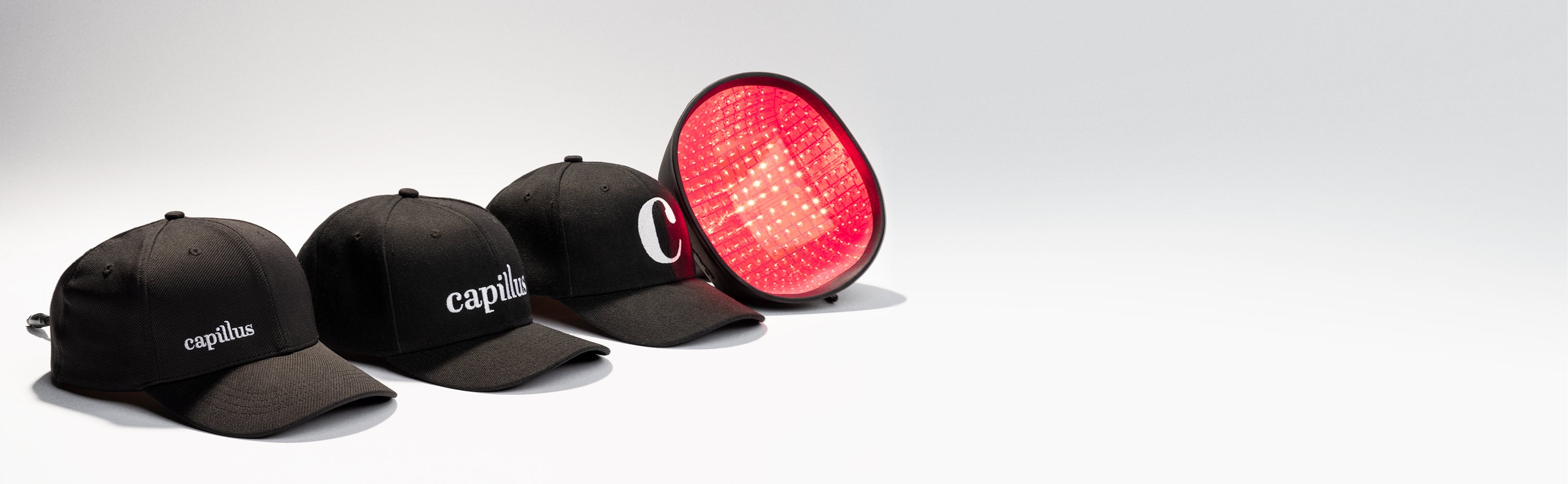 Three black baseball caps with “capillus” logos are lined up next to a red light therapy cap device that is glowing, all displayed on a white surface with a minimal background.