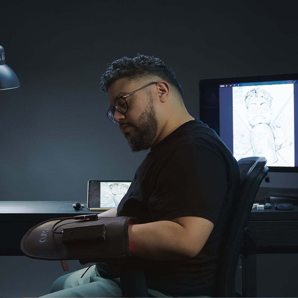 A man with a beard and glasses sits at a desk, wearing a padded glove on his arm. A tablet and a monitor displaying a sketch of a cartoon face are visible in the background, suggesting a creative workspace.