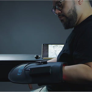 A man with a beard and glasses sits at a desk wearing a black therapy glove connected to a device. A tablet displaying a grayscale image is on the desk in front of him.