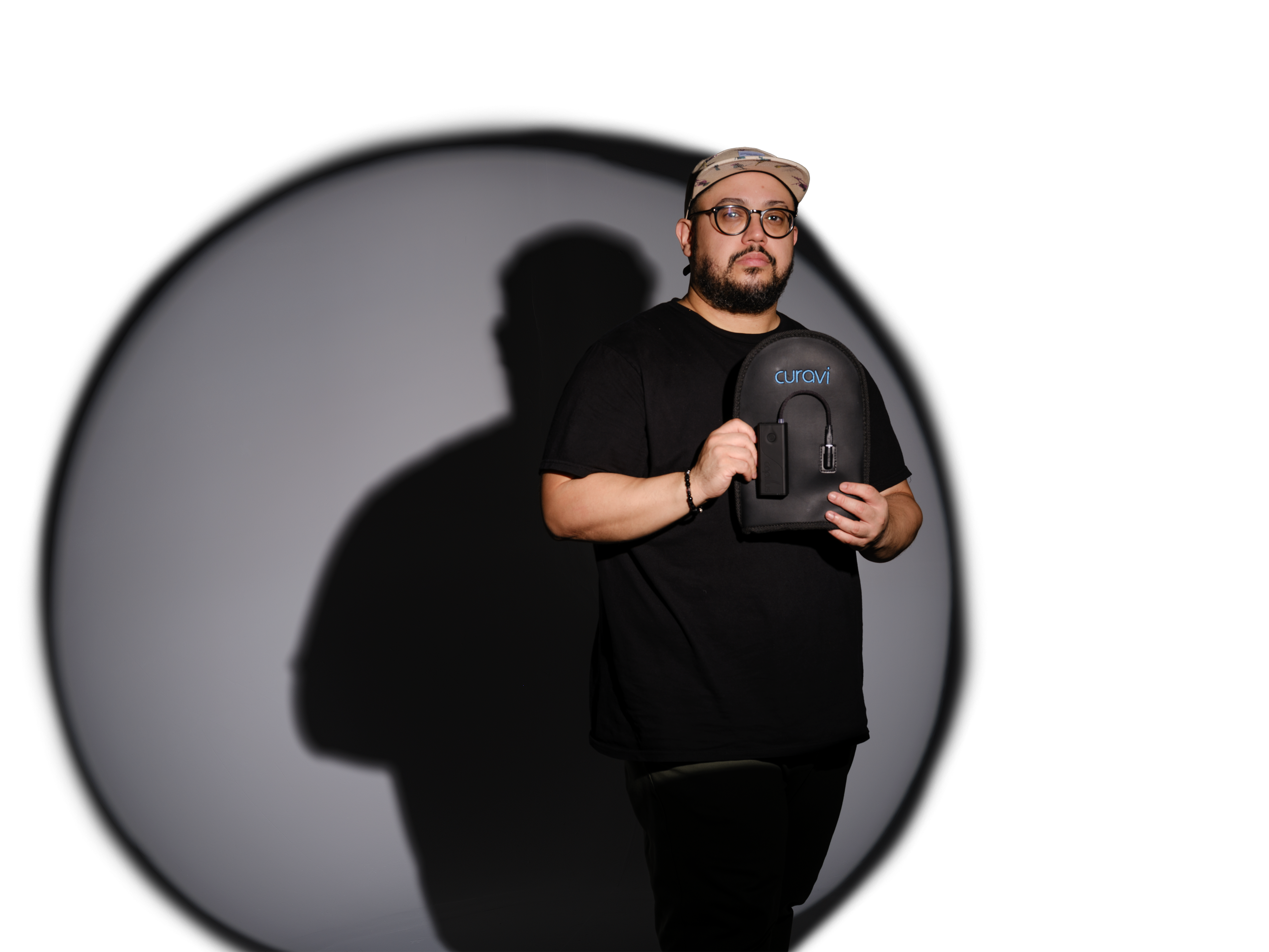 A person in a black outfit and glasses holds a device labeled “curaVIT” while standing in front of a spotlight on a white background. The person’s shadow is visible on the wall behind them.