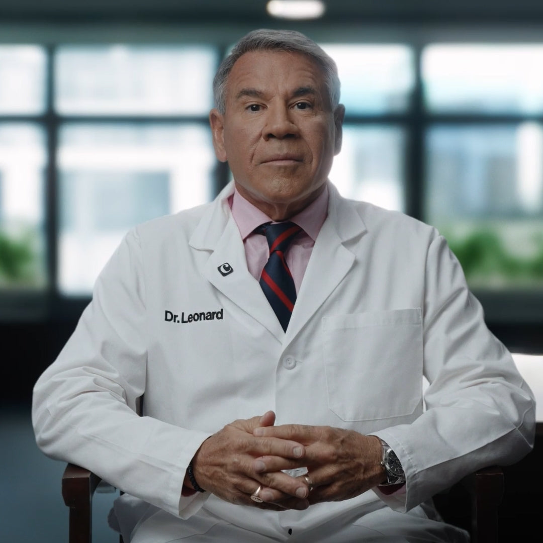 A mature man in a white lab coat labeled Dr. Leonard sits with hands clasped, facing the camera in a well-lit room with large windows and blurred greenery outside.