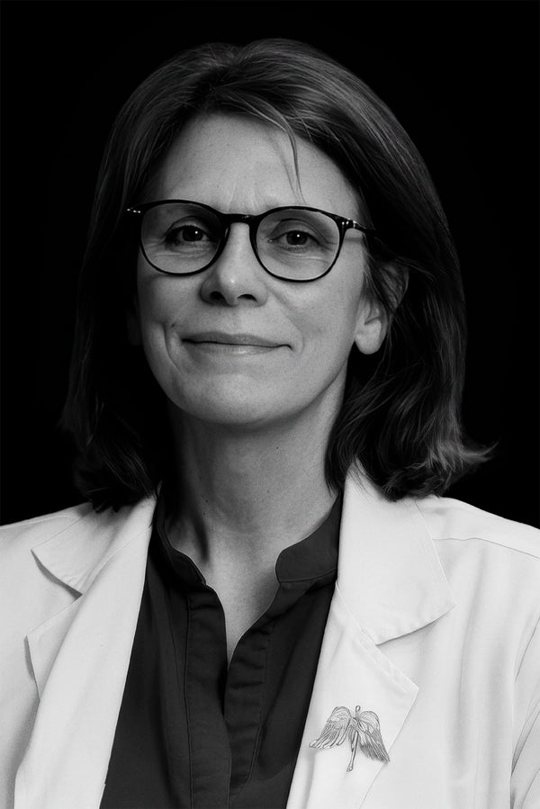 A woman with shoulder-length hair and glasses wears a white lab coat and a dark shirt, smiling slightly against a black background. A pin shaped like caduceus wings is visible on her lapel.