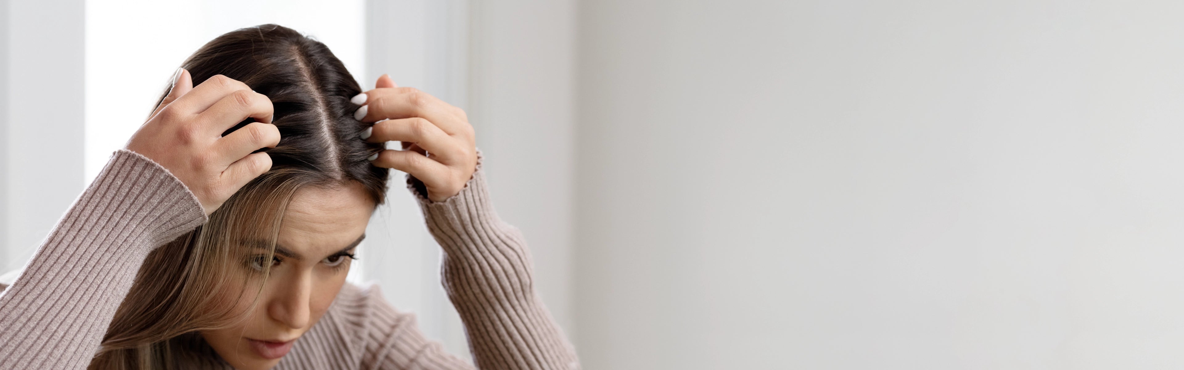 A person with long hair and a beige sweater examines their scalp closely with their hands, looking concerned, against a plain light background.