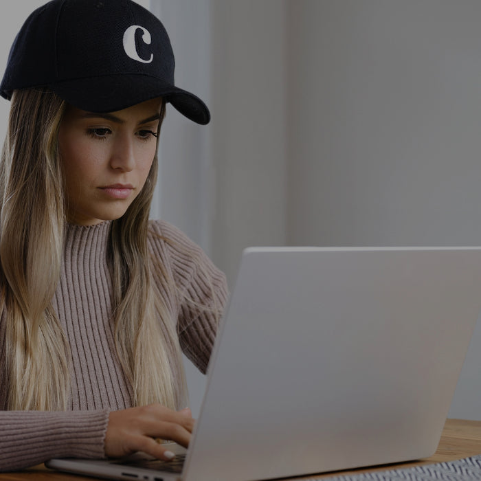 Une personne portant un chapeau noir orné d'un C blanc est assise à une table, devant un ordinateur portable. Elle a les cheveux longs et raides et est concentrée sur l'écran, dans une pièce bien éclairée.