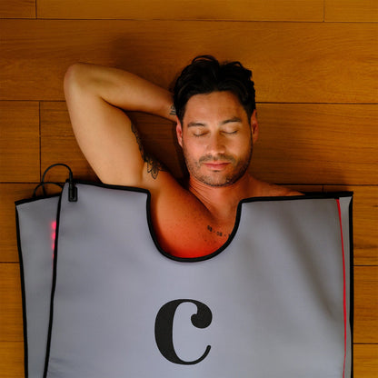 A man with dark hair relaxes with his eyes closed inside the gray Capillus Healing Pod sauna blanket by Capillus, featuring a large black C on the front—ideal for muscle recovery—set against a wooden wall background.