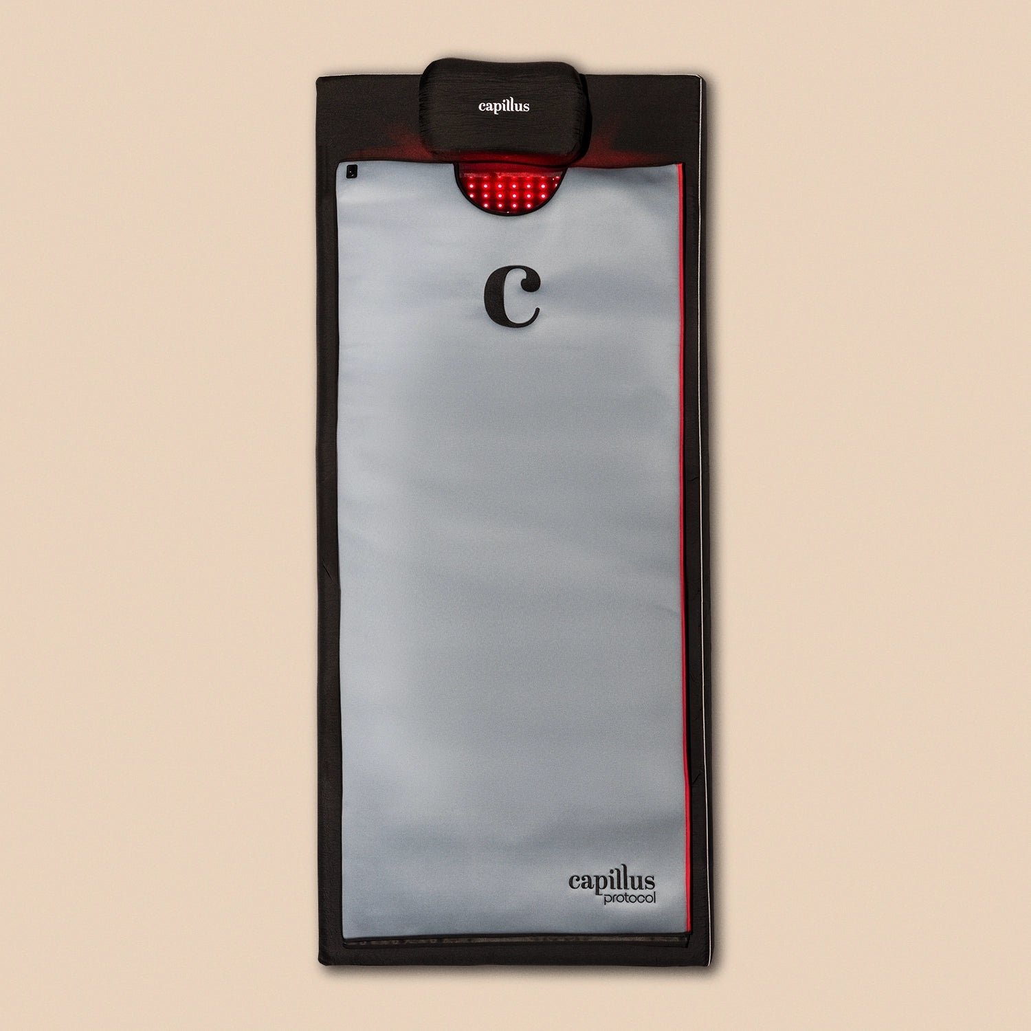 A rectangular gray and black Capillus Healing Pod by Capillus, featuring a red LED light panel at the top and labeled “capillus protocol,” designed for muscle recovery and joint pain relief, placed on a beige background.