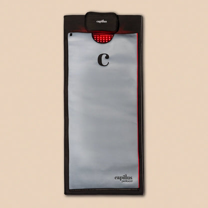 A rectangular gray and black Capillus Healing Pod by Capillus, featuring a red LED light panel at the top and labeled “capillus protocol,” designed for muscle recovery and joint pain relief, placed on a beige background.