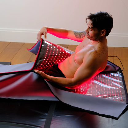 A shirtless man sits on the wooden floor, wrapping himself in the Capillus Healing Pod by Capillus—a large, flexible mat that emits red light for muscle recovery and wellness indoors.
