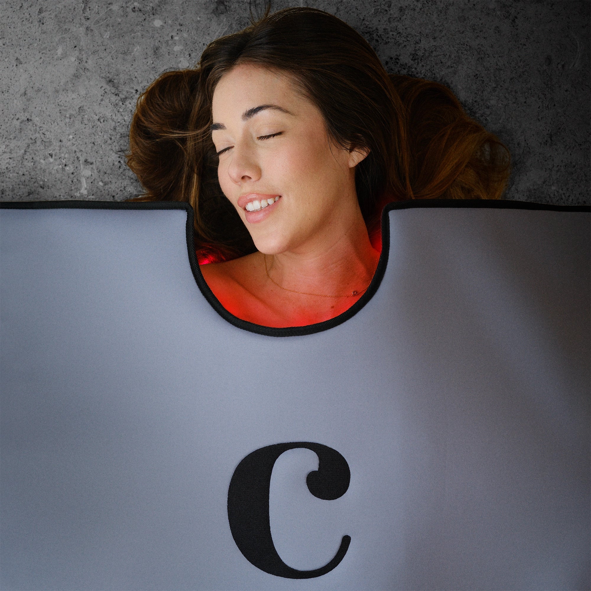 A woman with long brown hair relaxes on her back, eyes closed and smiling, wrapped in a gray Capillus Healing Pod by Capillus for muscle recovery on the concrete beneath her.