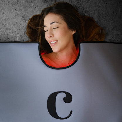 A woman with long brown hair relaxes on her back, eyes closed and smiling, wrapped in a gray Capillus Healing Pod by Capillus for muscle recovery on the concrete beneath her.