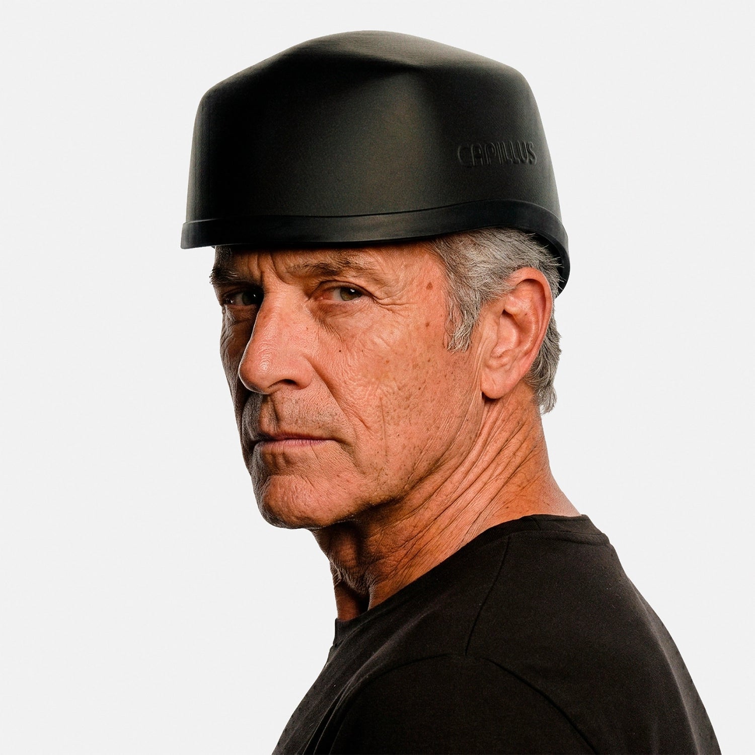 Un homme d'âge mûr, aux cheveux gris et au regard sérieux, porte le casque Capillus Neuro noir mat de la marque Capillus, conçu pour la luminothérapie rouge et proche infrarouge, tout en posant devant un fond blanc uni.