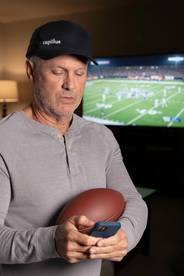 A man wearing a cap holds a football and looks at his smartphone. A football game is playing on the TV in the background. The room is dimly lit with a lamp beside the TV.