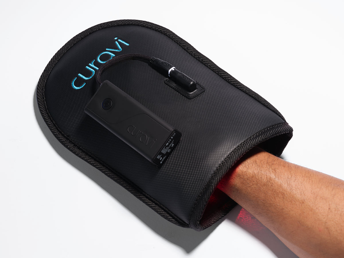 A persons arm is inserted into a black Curavi device with a visible control panel, designed for therapy or treatment, lying on a white surface.