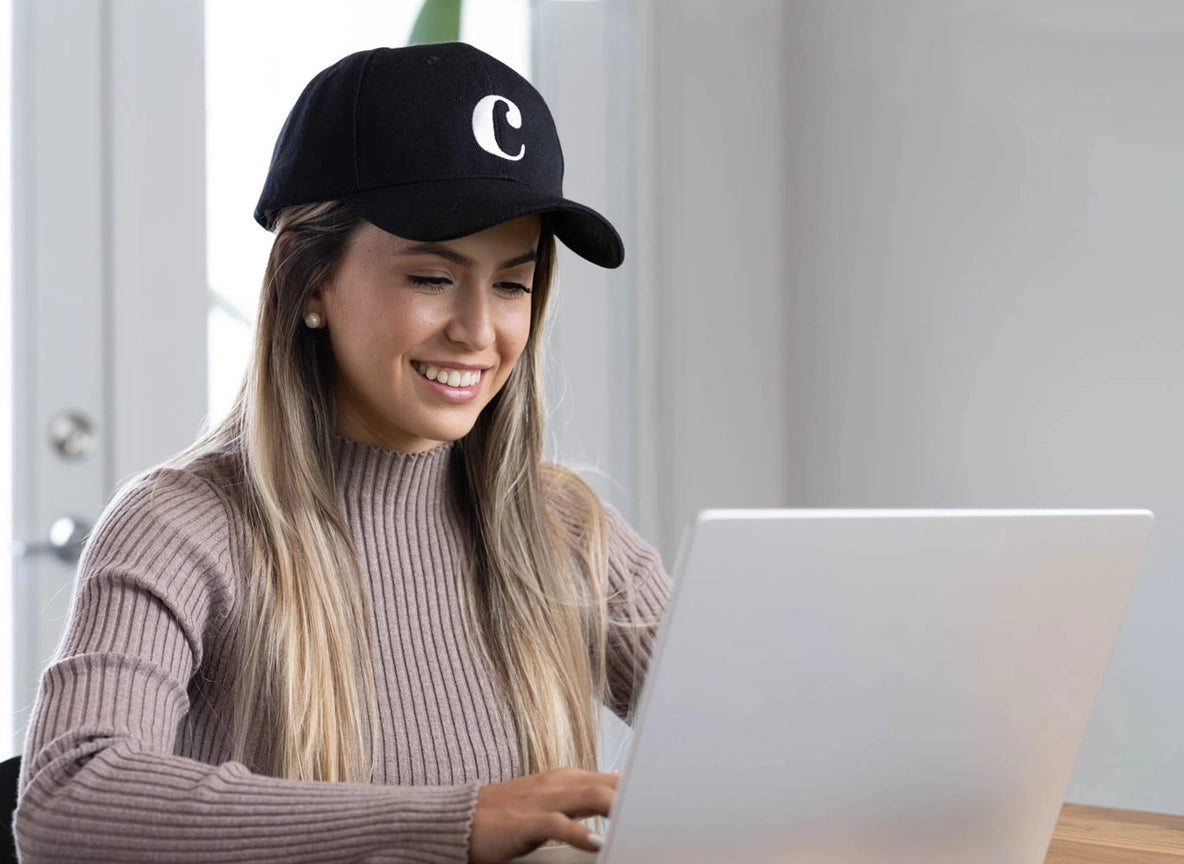 A smiling woman wearing a black cap with the letter C on it and a beige sweater is sitting at a table, working on a laptop in a bright room.